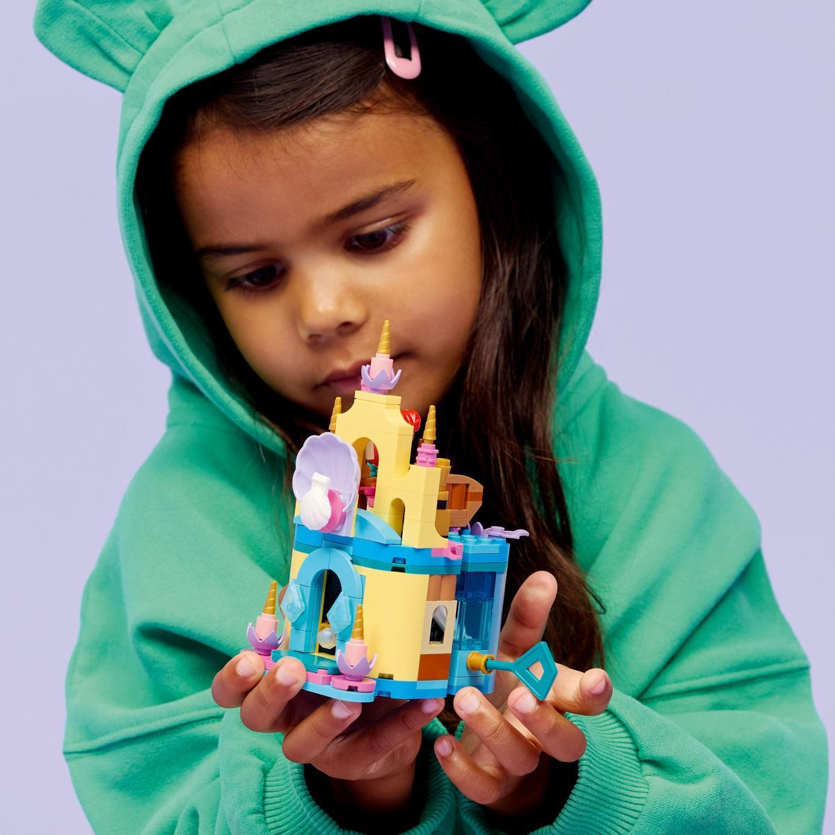 Una niña con una sudadera con capucha verde sostiene un castillo de juguete colorido. El castillo tiene elementos amarillos, azules y rosas.