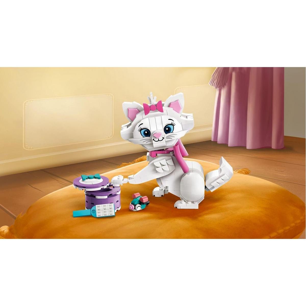 Biały kot Lego z różową kokardą i szalikiem siedzi na żółtej poduszce. Obok zabawki Lego i różowa zasłona.