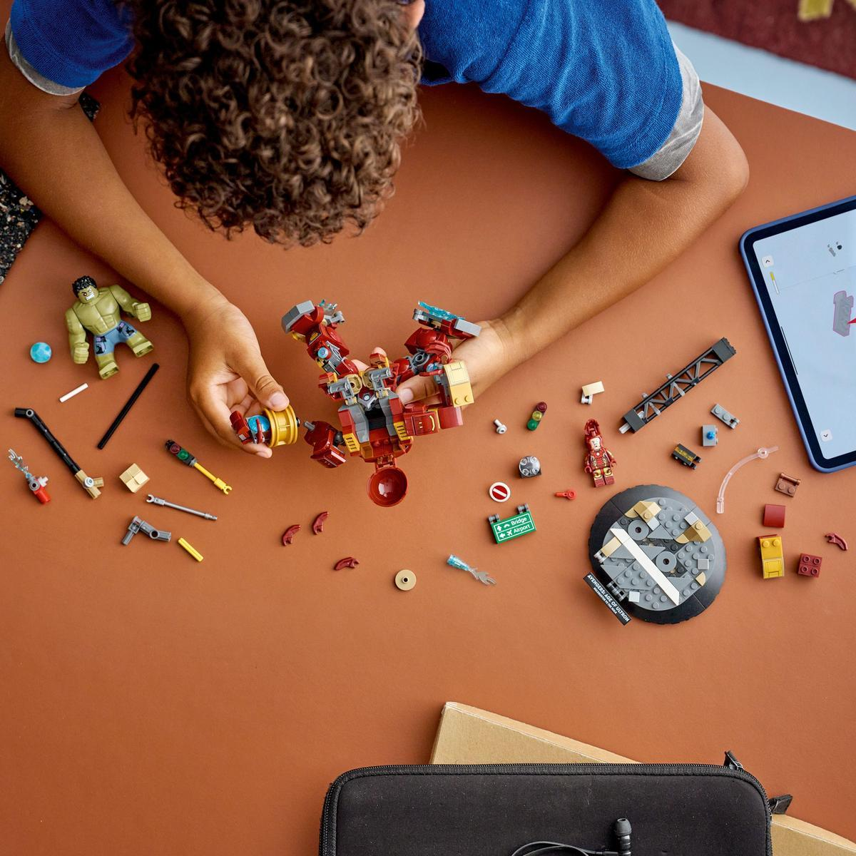 Dziecko składa zestaw Lego Iron Man na brązowej powierzchni, otoczone elementami, tabletem i figurką Hulka.