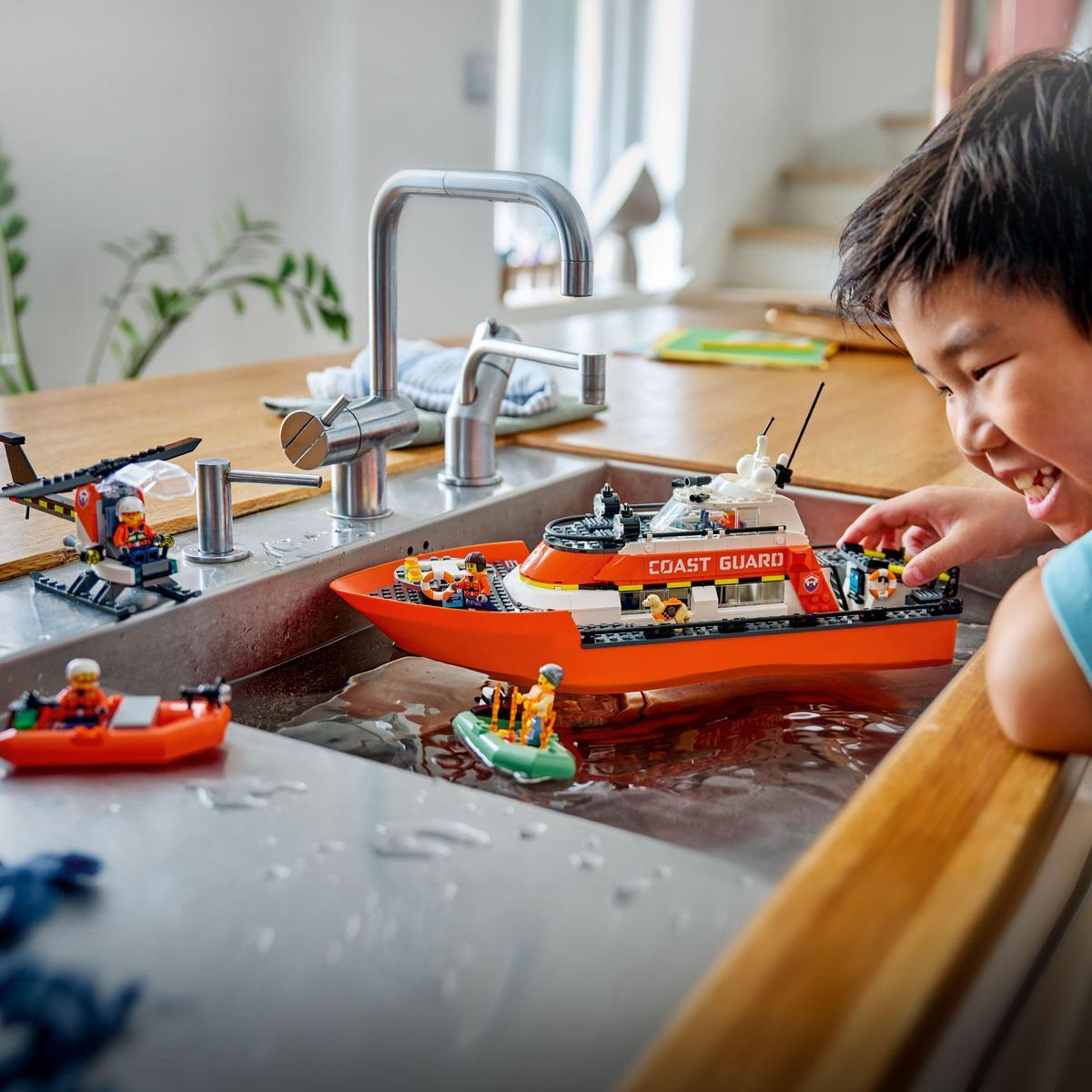 Dziecko bawi się zabawkami Lego Straży Przybrzeżnej w zlewie wypełnionym wodą. Obecne są łódź, helikopter i ponton.