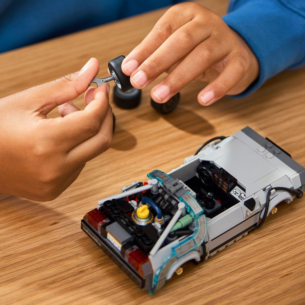 Osoba składa samochód Lego DeLorean. Samochód jest szary, z czarnym wnętrzem. Osoba trzyma koło i narzędzie.