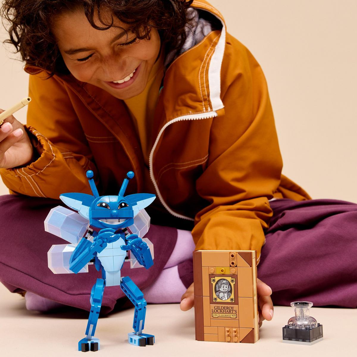 Uśmiechnięte dziecko siedzi z niebieskim stworzeniem Lego, książką i kałamarzem. Dziecko ma na sobie brązową kurtkę i fioletowe spodnie.