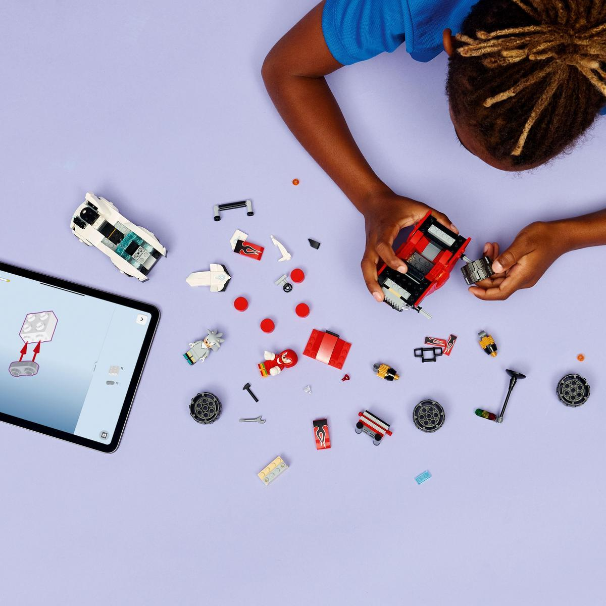 Dziecko składa czerwoną zabawkę samochodzik. Różne części samochodzika i tablet z instrukcjami leżą na fioletowej powierzchni.