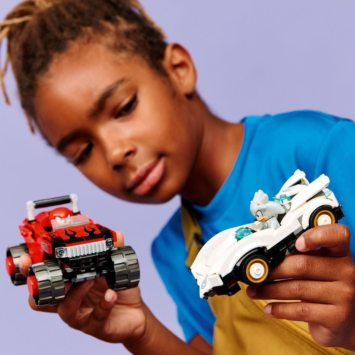 Chłopiec trzyma dwa zabawki samochody, czerwony i biały z postacią w środku. Ma na sobie niebieską koszulę, a tło jest fioletowe.