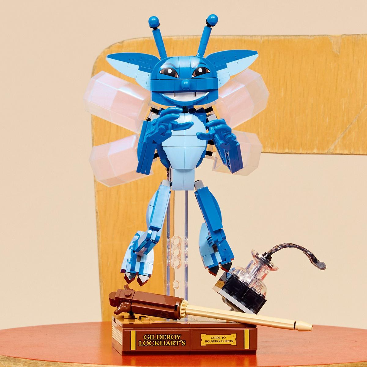 Niebieska postać z klocków Lego ze skrzydłami, stojąca na książce. Obok pióro i kałamarz. Tłem jest drewniane krzesło.