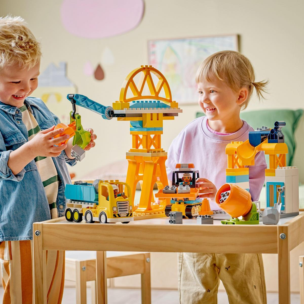 Dwoje dzieci bawi się zabawkami konstrukcyjnymi. Na stole stoi dźwig, ciężarówka i mieszalnik. Oboje dzieci uśmiechają się radośnie.