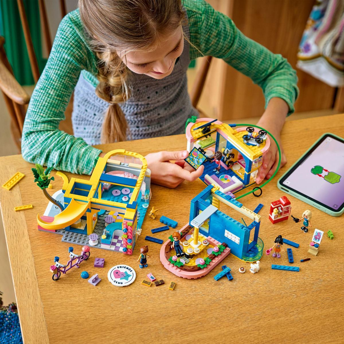 Una ragazza gioca con set LEGO Friends su un tavolo di legno. Ci sono più strutture e accessori. Un tablet mostra le istruzioni.