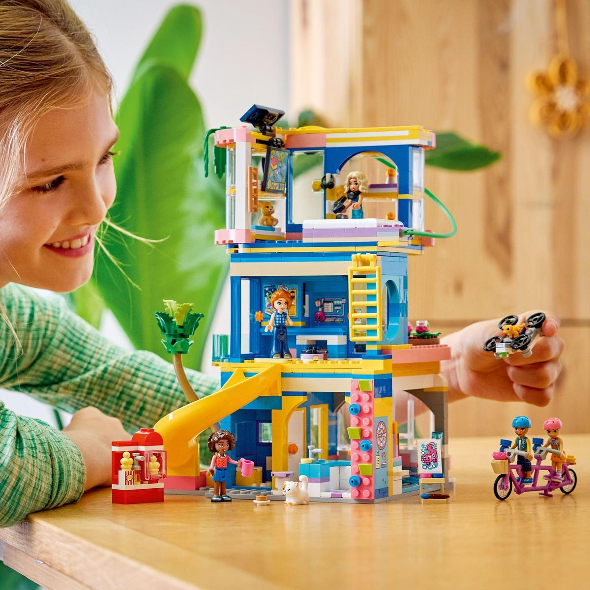Una ragazza sorride a un set Lego. Il set mostra un edificio con personaggi e accessori. Un'altra persona tiene un drone.