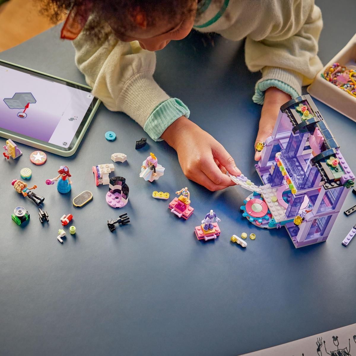 Dziecko buduje zestaw Lego, z tabletem pokazującym instrukcje. Różne elementy Lego są rozrzucone na ciemnoniebieskiej powierzchni.