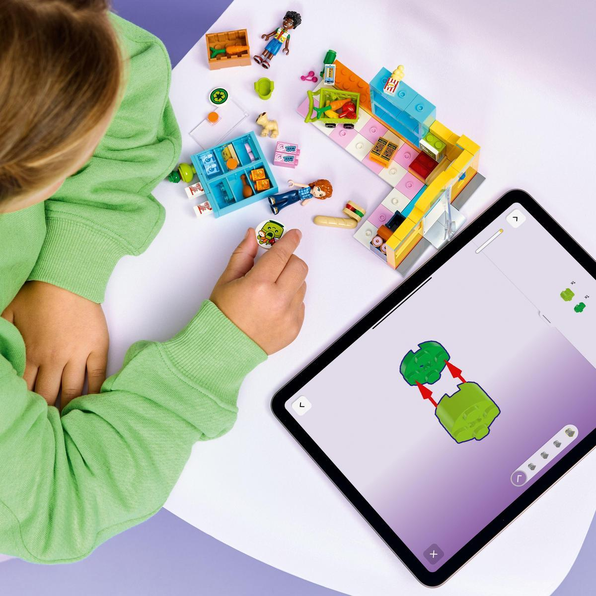 Yeşil bir kazaklı çocuk Lego oynuyor. Bir tablet talimatları gösteriyor. Lego figürleri ve yiyecekler masaya dağılmış durumda.