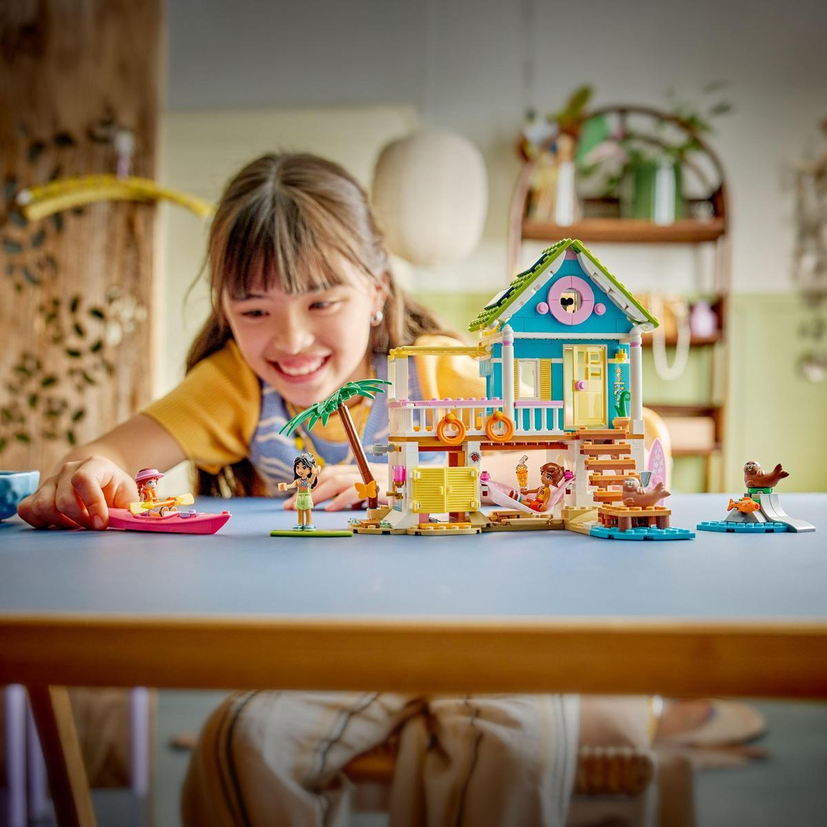 Dziewczyna uśmiecha się do zestawu LEGO z domkiem na plaży z postaciami w łodzi, na plaży i odpoczywającymi. Zestaw jest na niebieskim stole.