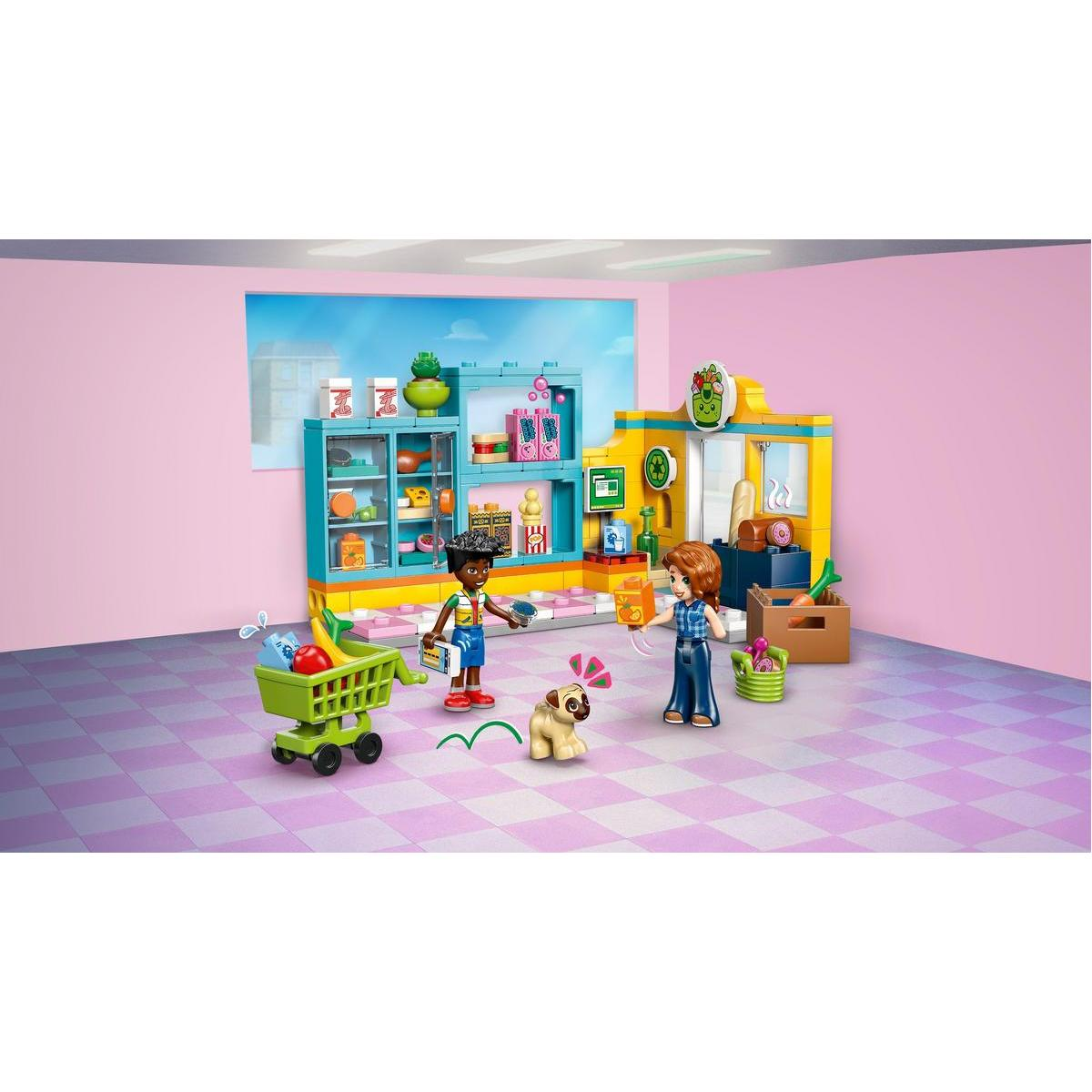İki figür, bir köpek, meyve dolu bir sepet ve raflarla dolu bir Lego süpermarket sahnesi. Pembe duvarlar ve fayanslı zemin.