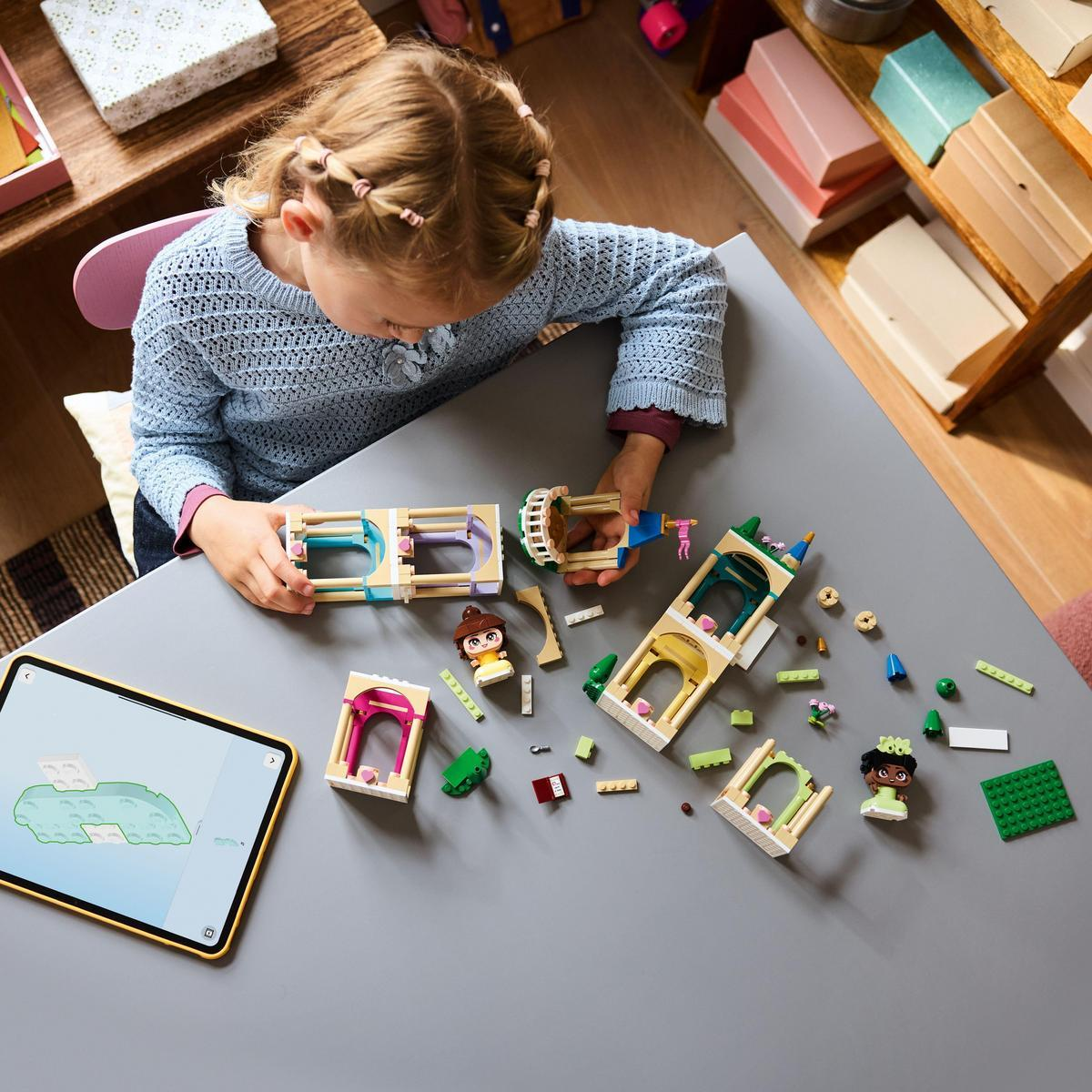 Dziewczynka buduje zestaw Lego na szarym stole. Klocki Lego i tablet z instrukcjami są rozrzucone.