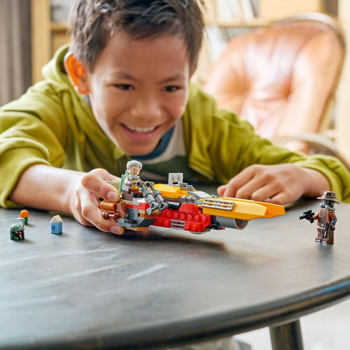 Bir çocuk, bir masanın üzerindeki bir LEGO setine gülümsüyor. Set, sarı ve kırmızı bir araç, iki minifigür ve diğer küçük parçaları içerir.