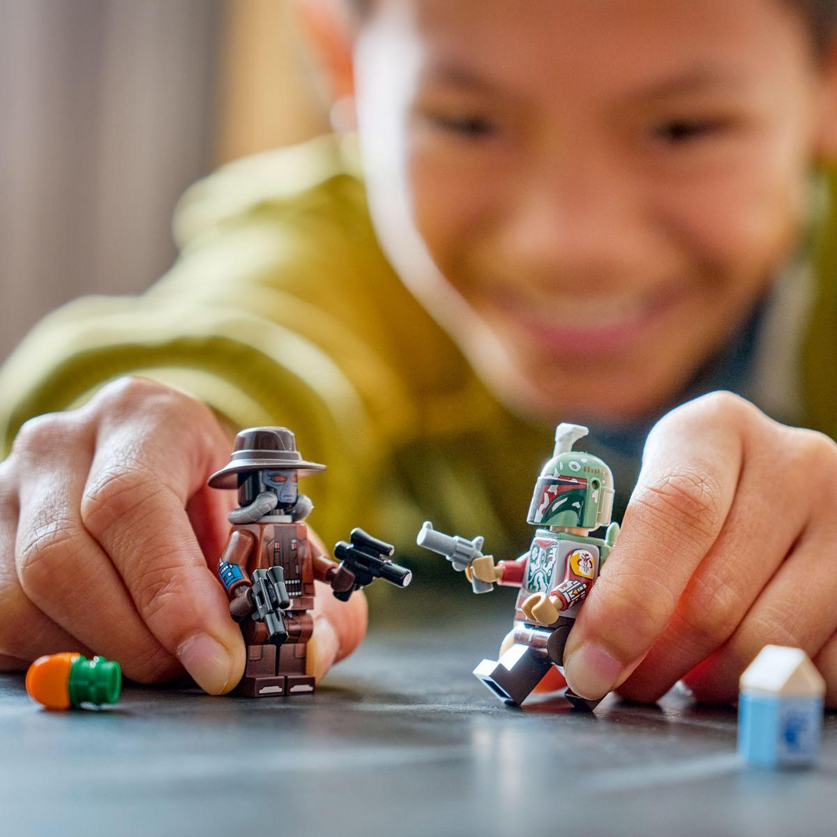 Bir çocuk Lego Star Wars figürleriyle oynuyor. Bir figür kahverengi şapka, diğeri yeşil miğfer takıyor. Her ikisi de blaster tutuyor.