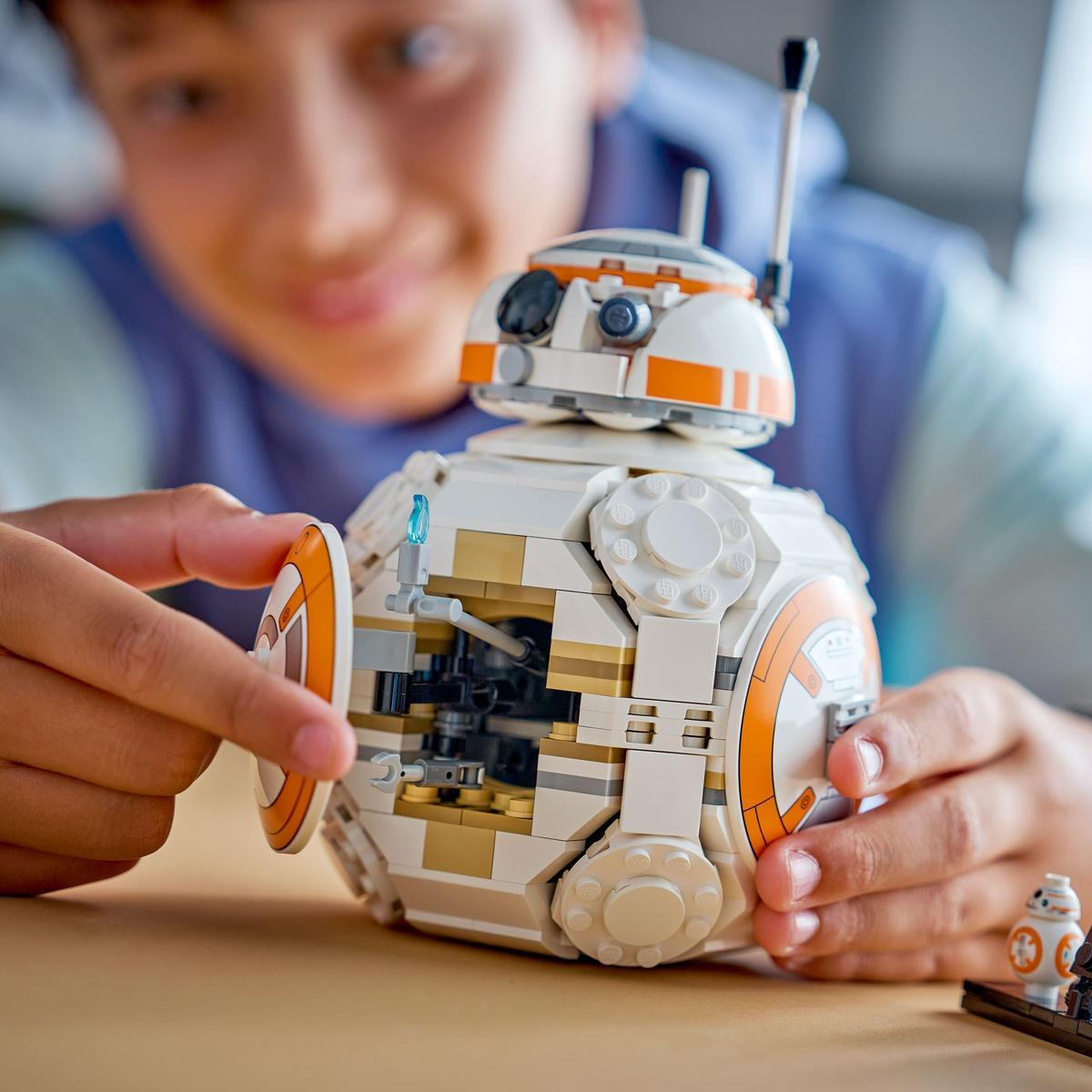 Chłopiec trzyma zestaw Lego BB-8. Biało-pomarańczowy robot jest częściowo zmontowany. Małe Lego BB-8 leży na stole.