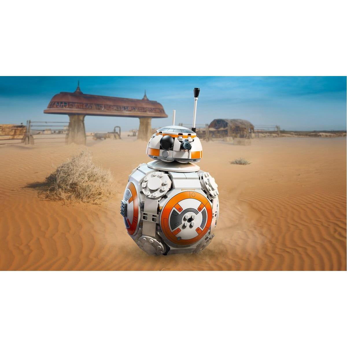 Zabawka robota BB-8 w scenerii pustynnej z budynkiem i piaskiem. Robot jest biało-pomarańczowy.