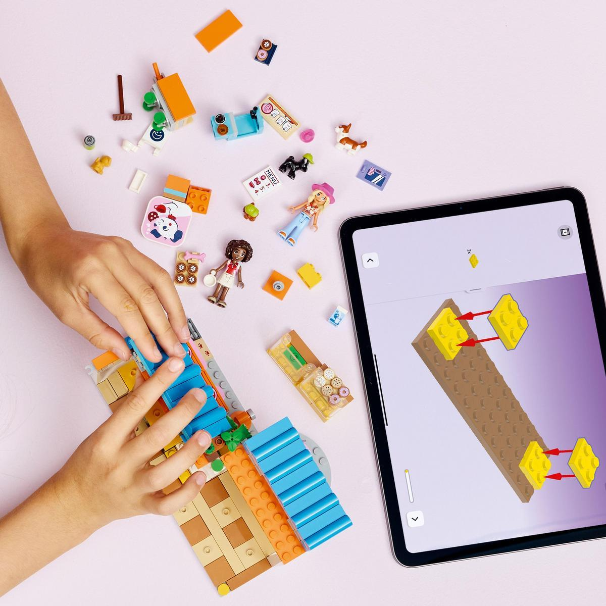 Ręce składające zestaw Lego. Tablet pokazuje instrukcje. Różne klocki i figurki Lego rozrzucone na różowej powierzchni.