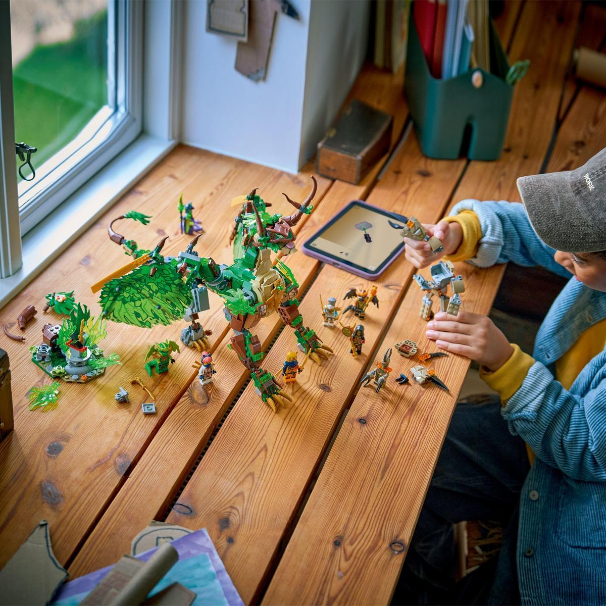 Dziecko bawi się smokami i figurkami Lego na drewnianym stole. Tablet pokazuje instrukcje. Scena rozgrywa się blisko okna.