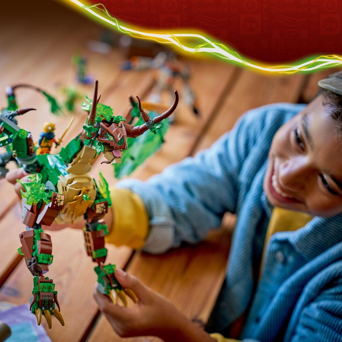 Uśmiechnięta osoba trzyma zielony zestaw Lego z smokiem. Smok jest na drewnianym stole. Inne zestawy Lego są w tle.