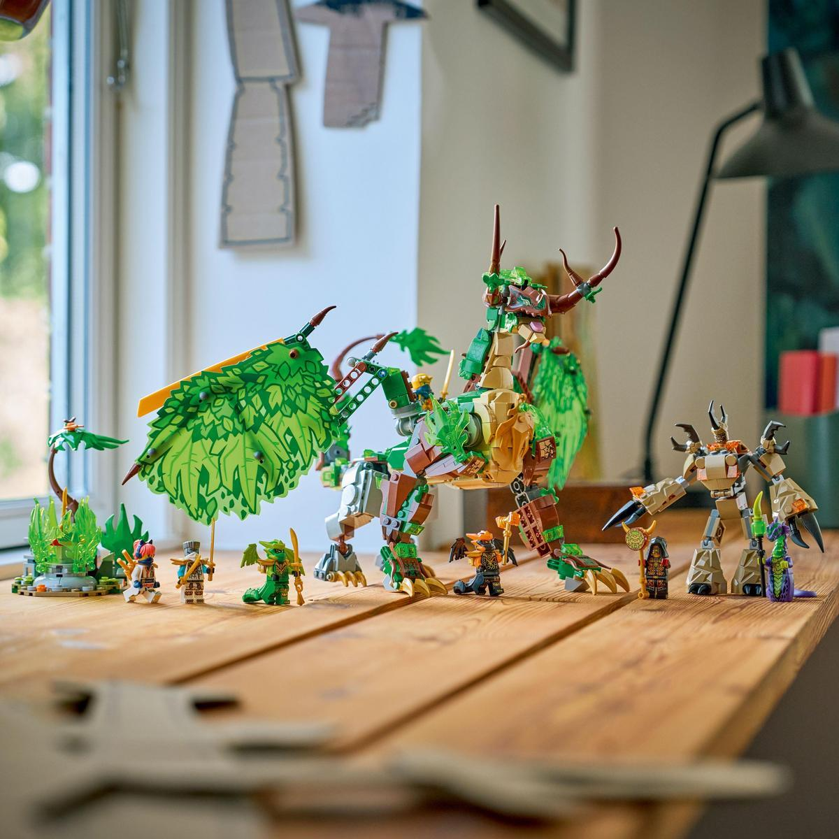 Na drewnianym stole widnieje smok Lego, figurki i inne konstrukcje Lego. Smok ma zielone i brązowe elementy. Figurki stoją w rzędzie.