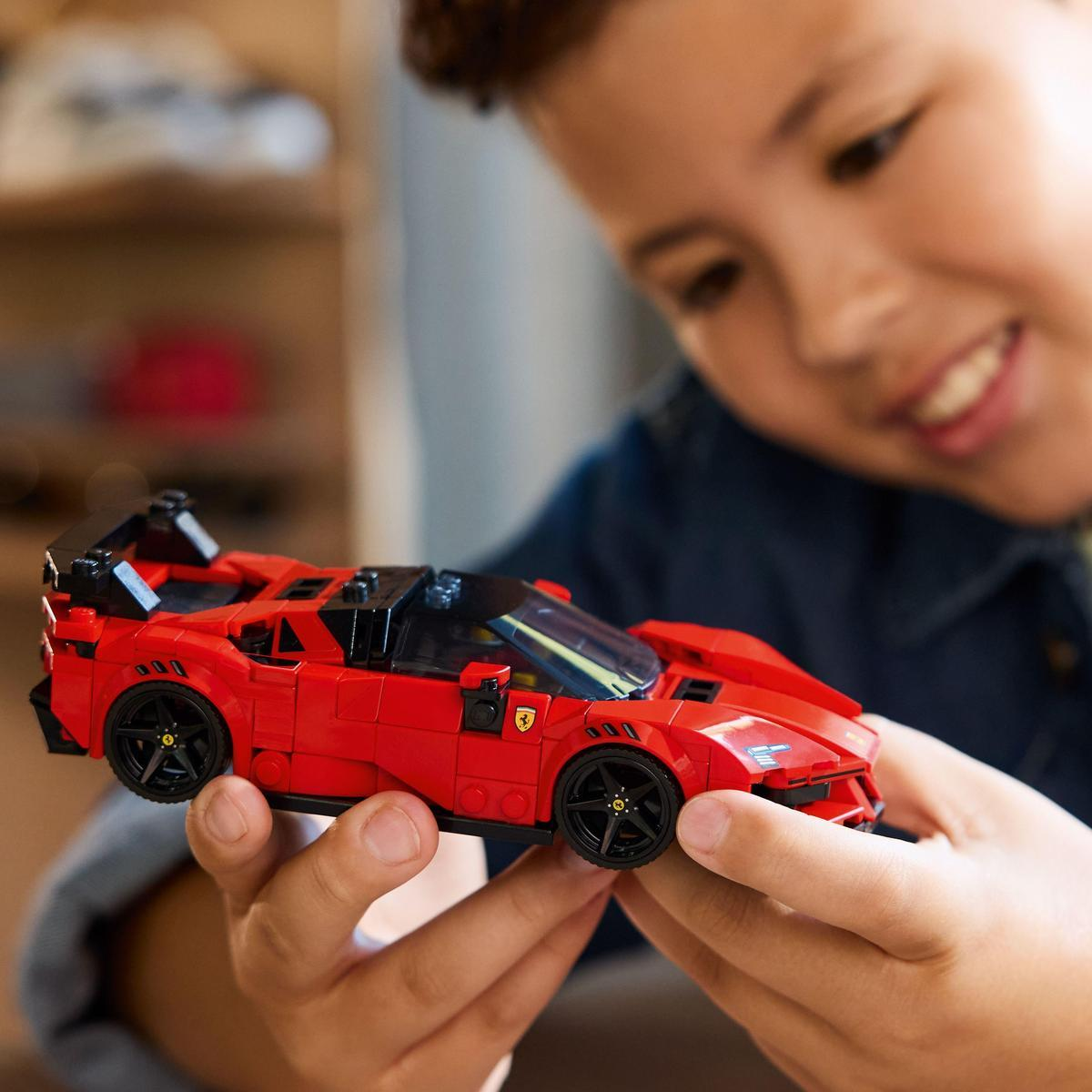 Uśmiechnięte dziecko trzyma czerwone Lego Ferrari. Samochód ma czarne koła i akcenty. Tło jest rozmyte.