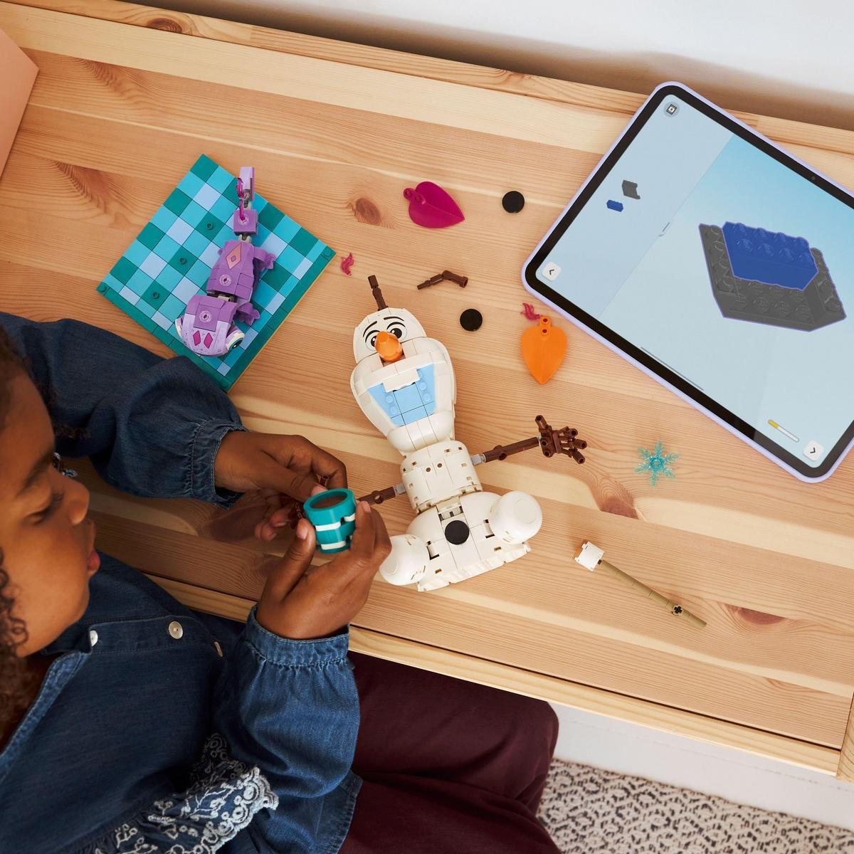 Dziecko składa zestaw Lego Olafa, z tabletem pokazującym instrukcje. Części i inne klocki Lego są rozrzucone na drewnianym stole.
