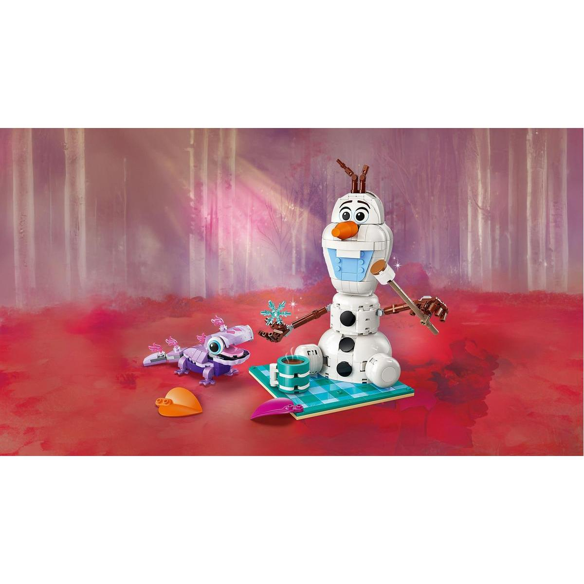 Olaf i fioletowa zabawka smoka leżą na turkusowej macie. Tło to różowy las.