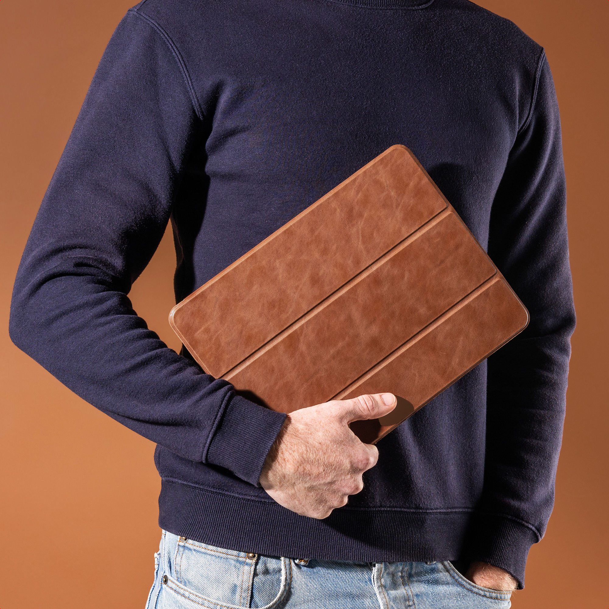Un homme en pull-over bleu marine et jean tient un étui marron pour tablette sur fond marron.