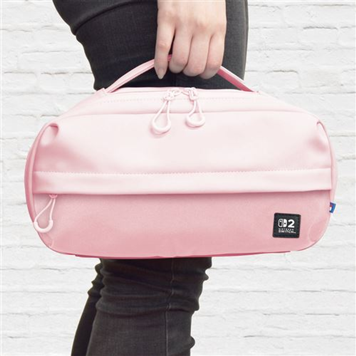 Una persona sostiene una funda de transporte rosa para Switch. La funda tiene dos asas, cremalleras y un pequeño logotipo. El fondo es una pared de ladrillos blancos.