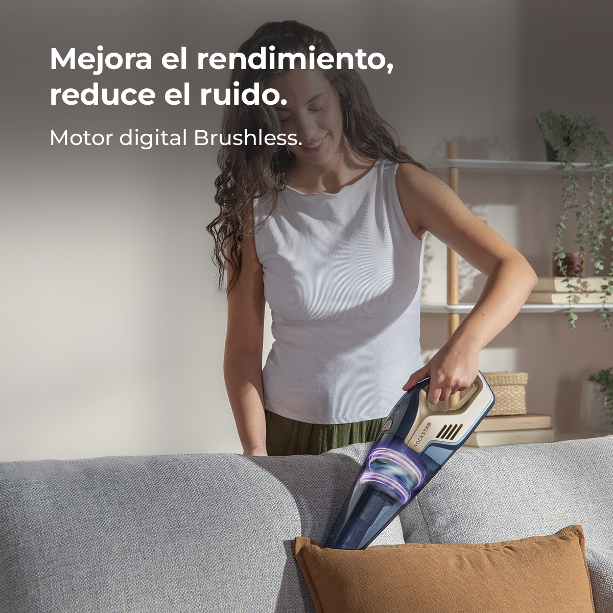 Una mujer aspira un sofá con una aspiradora de mano. La aspiradora es azul y beige. La mujer usa top blanco y pantalones verdes.