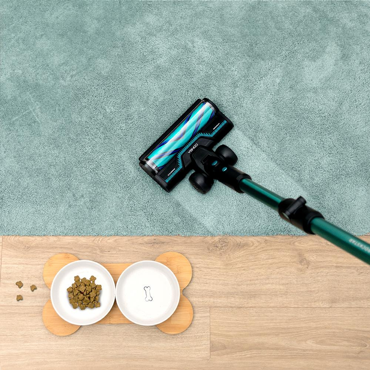 Un aspirapolvere sta pulendo un tappeto verde acqua. Le ciotole per cani sono sul pavimento in legno. Un po' di cibo è sul pavimento.