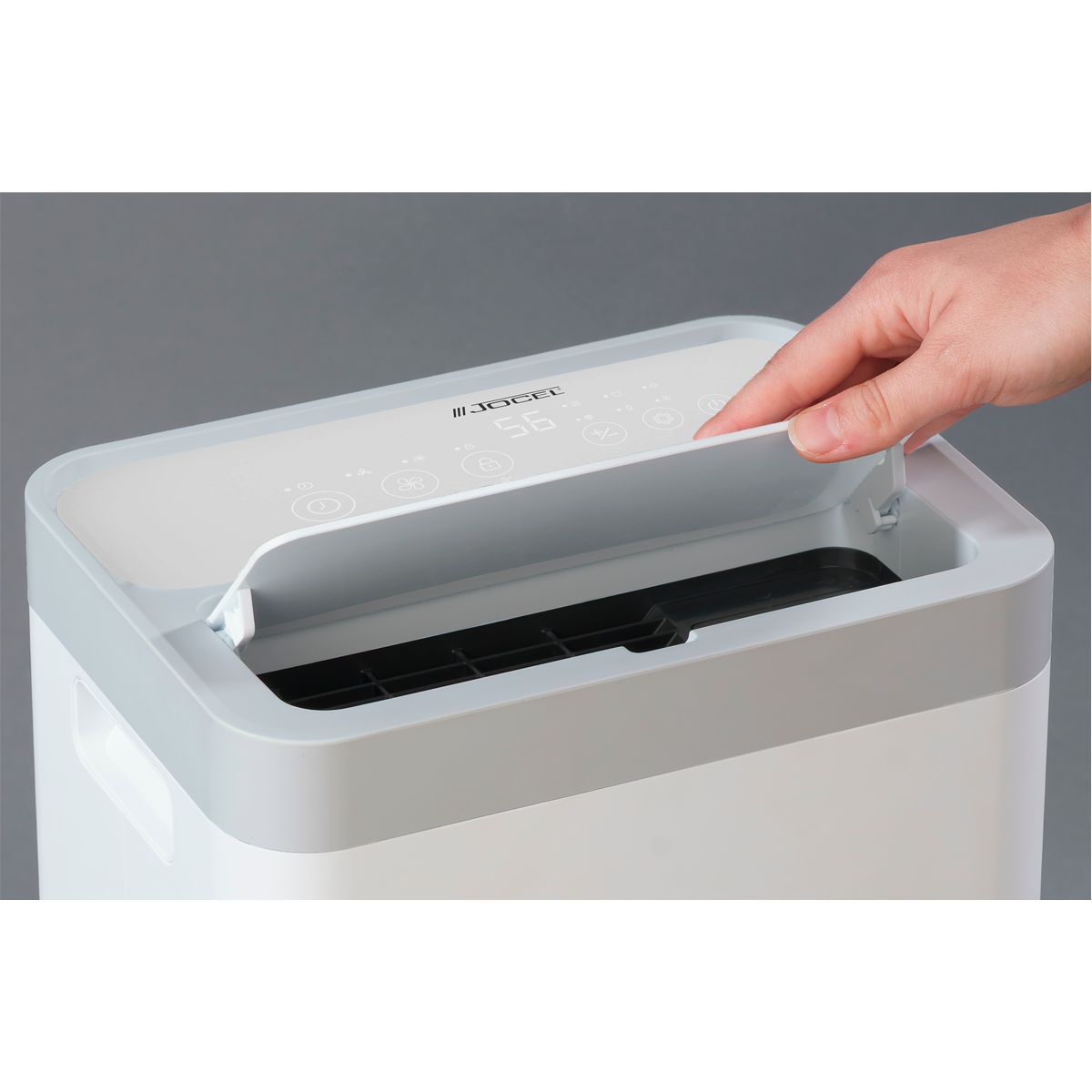 Una mano apre il coperchio di un purificatore d'aria bianco. La parte superiore ha un display digitale con '56'. Lo sfondo è grigio.