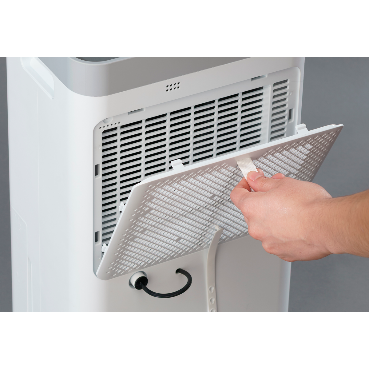 Una mano estrae un filtro da un purificatore d'aria bianco. Il purificatore ha una griglia nera e uno sfondo grigio.