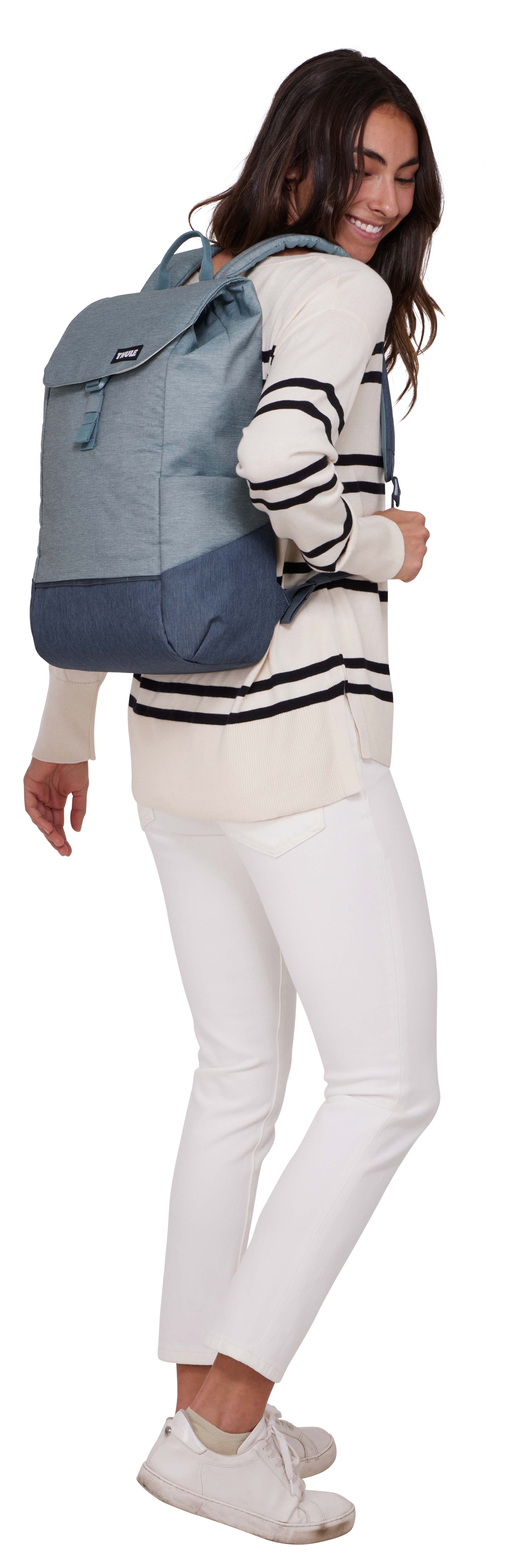 Eine Frau trägt einen gestreiften Pullover und eine weiße Hose, trägt einen blauen Rucksack und lächelt.
