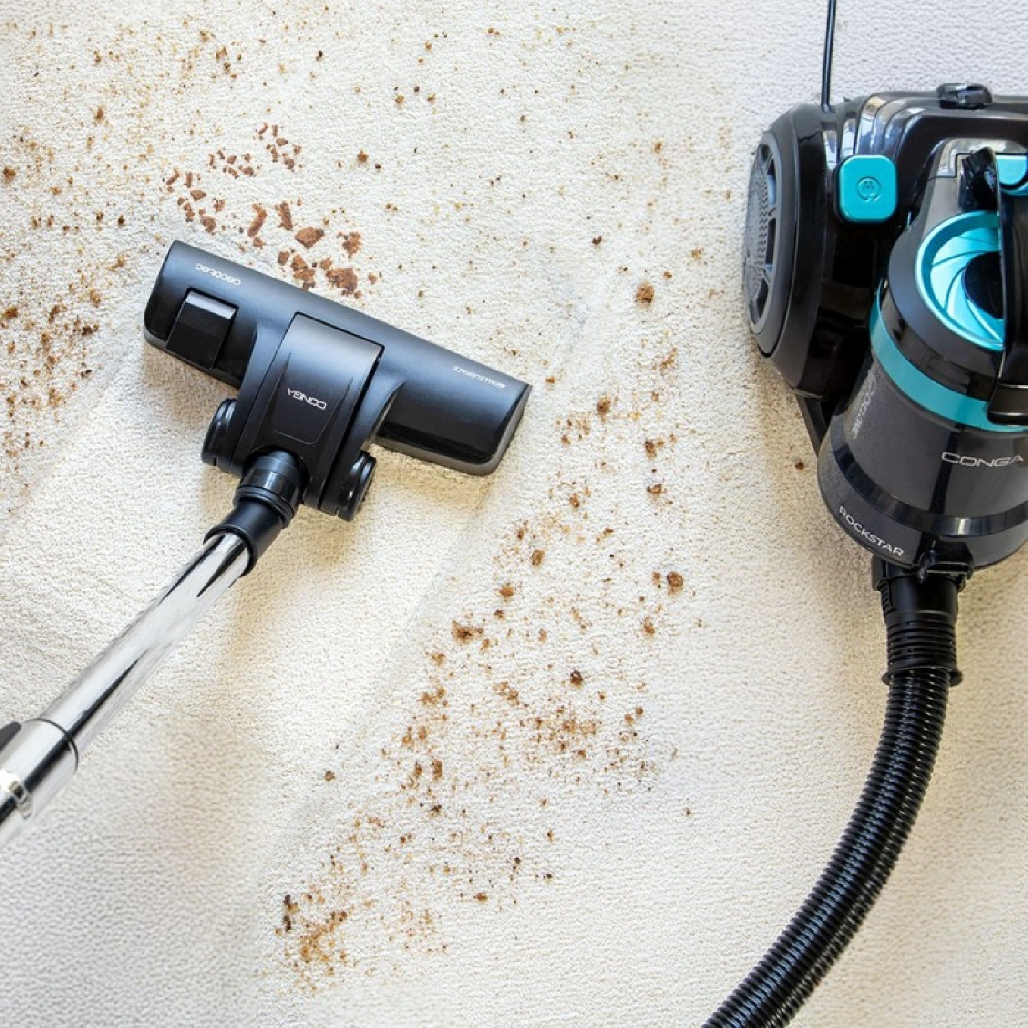 Una testina di aspirapolvere nera pulisce un tappeto chiaro con macchie marroni. Sullo sfondo un aspirapolvere nero e turchese.