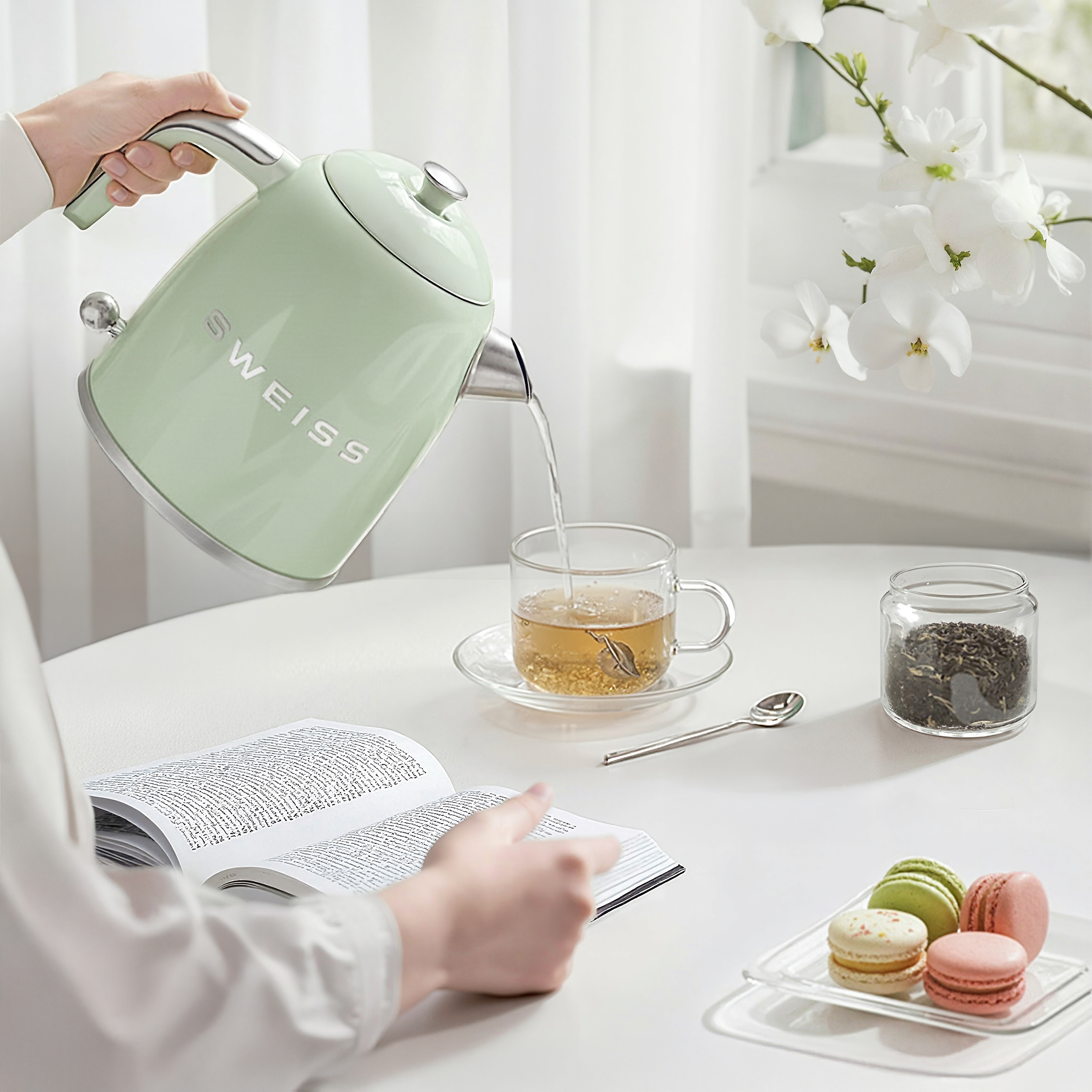 Eine Person gießt heißes Wasser aus einem grünen Wasserkocher in eine Glastasse. Bücher, Makronen und Blumen liegen auf dem Tisch.