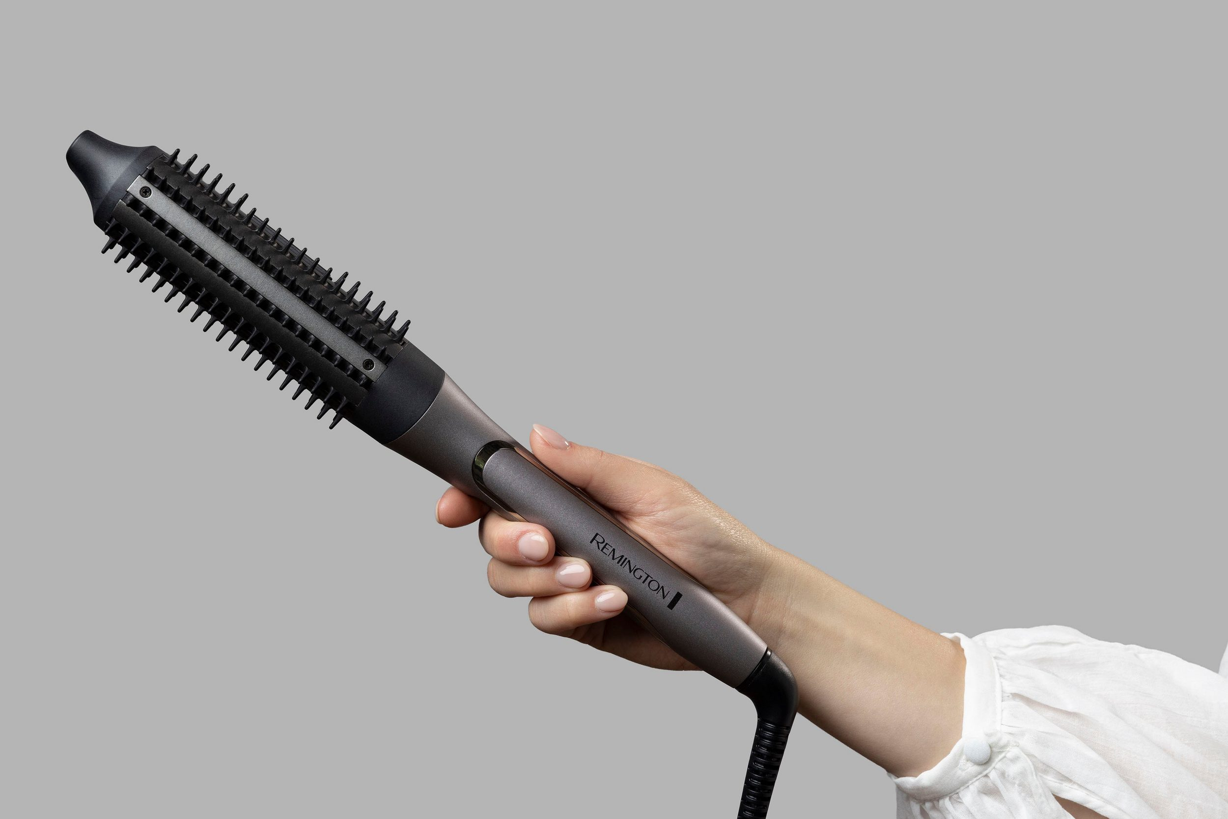 Bir el, gri bir arka plan üzerinde gri bir Remington saç şekillendirme fırçası tutuyor. Fırça siyah ve gridir.