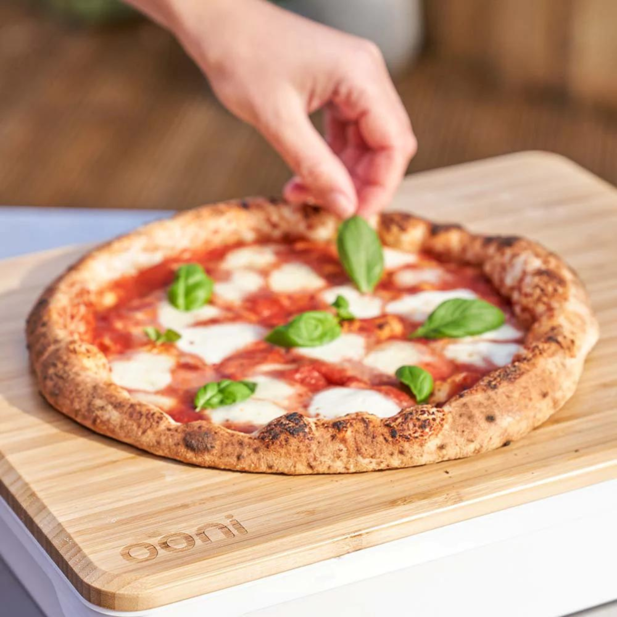 Eine Hand legt Basilikumblätter auf eine Pizza. Die Pizza hat Tomatensauce, Mozzarella und liegt auf einem Holzbrett.