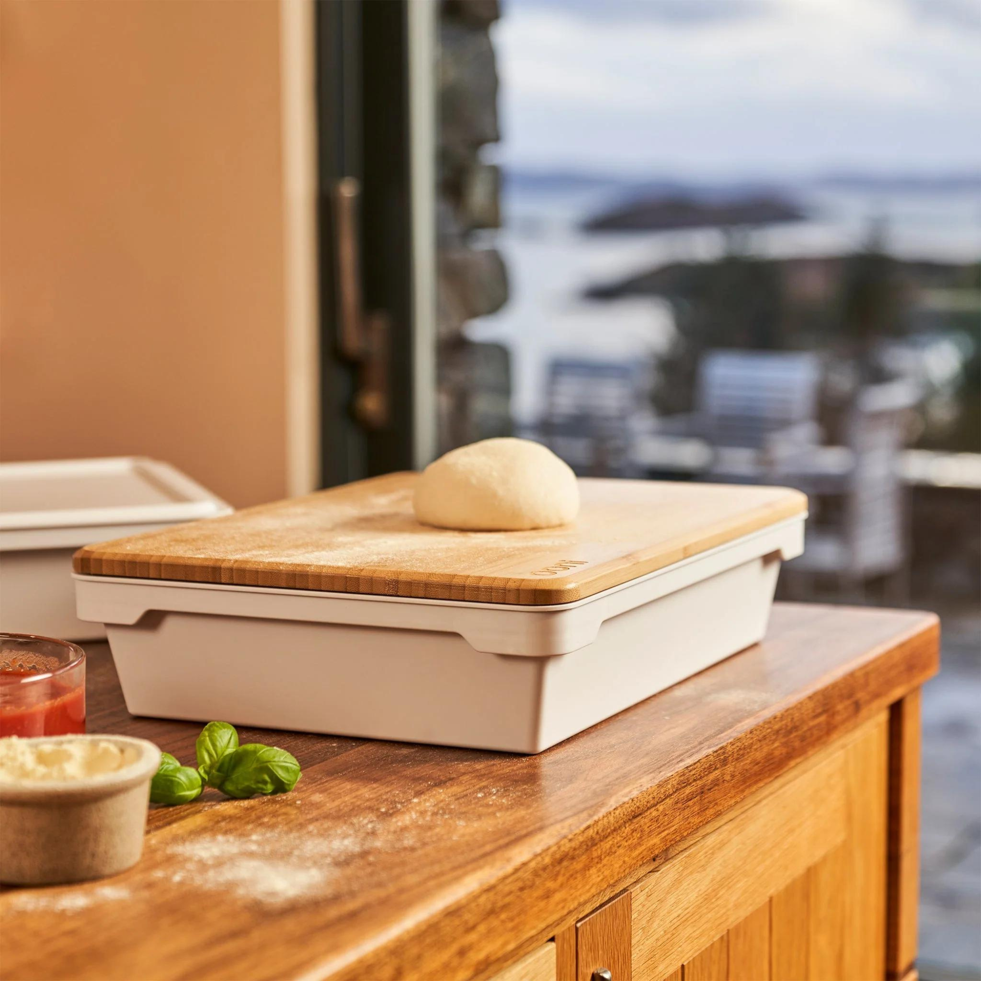 Ein Holztisch hält ein teigbedecktes Tablett mit einem Teigball, Basilikum, Sauce und Mehl. Ein Fenster zeigt eine verschwommene Landschaft.