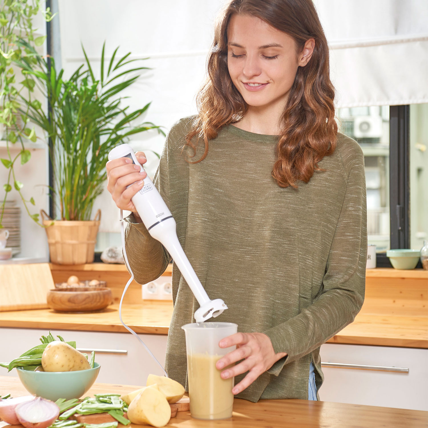 Kobieta w zielonej koszuli blenduje zupę w kuchni. Składniki, takie jak ziemniaki i cebula, są na stole. Rośliny i okno w tle.