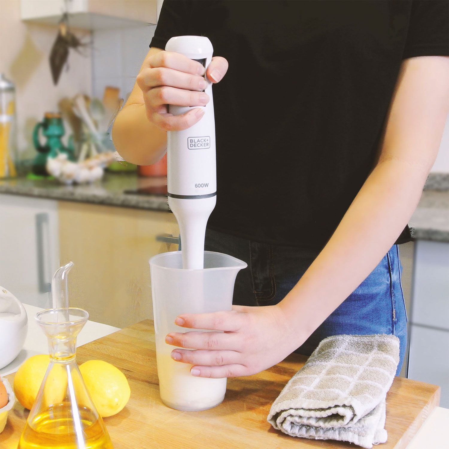 Osoba używa blendera ręcznego Black+Decker do mieszania składników w miarce. Na blacie znajdują się również cytryny, olej i ręcznik.