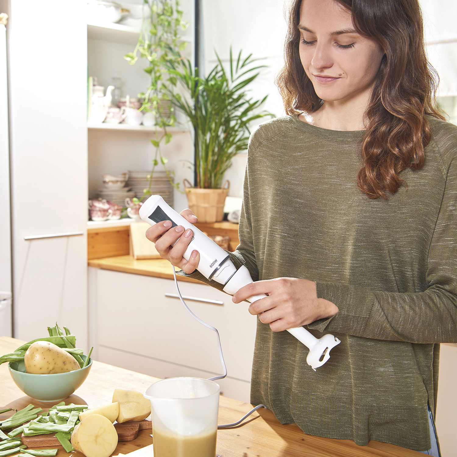 Kobieta w zielonym swetrze trzyma biały blender ręczny w kuchni. Warzywa i zupa na stole.