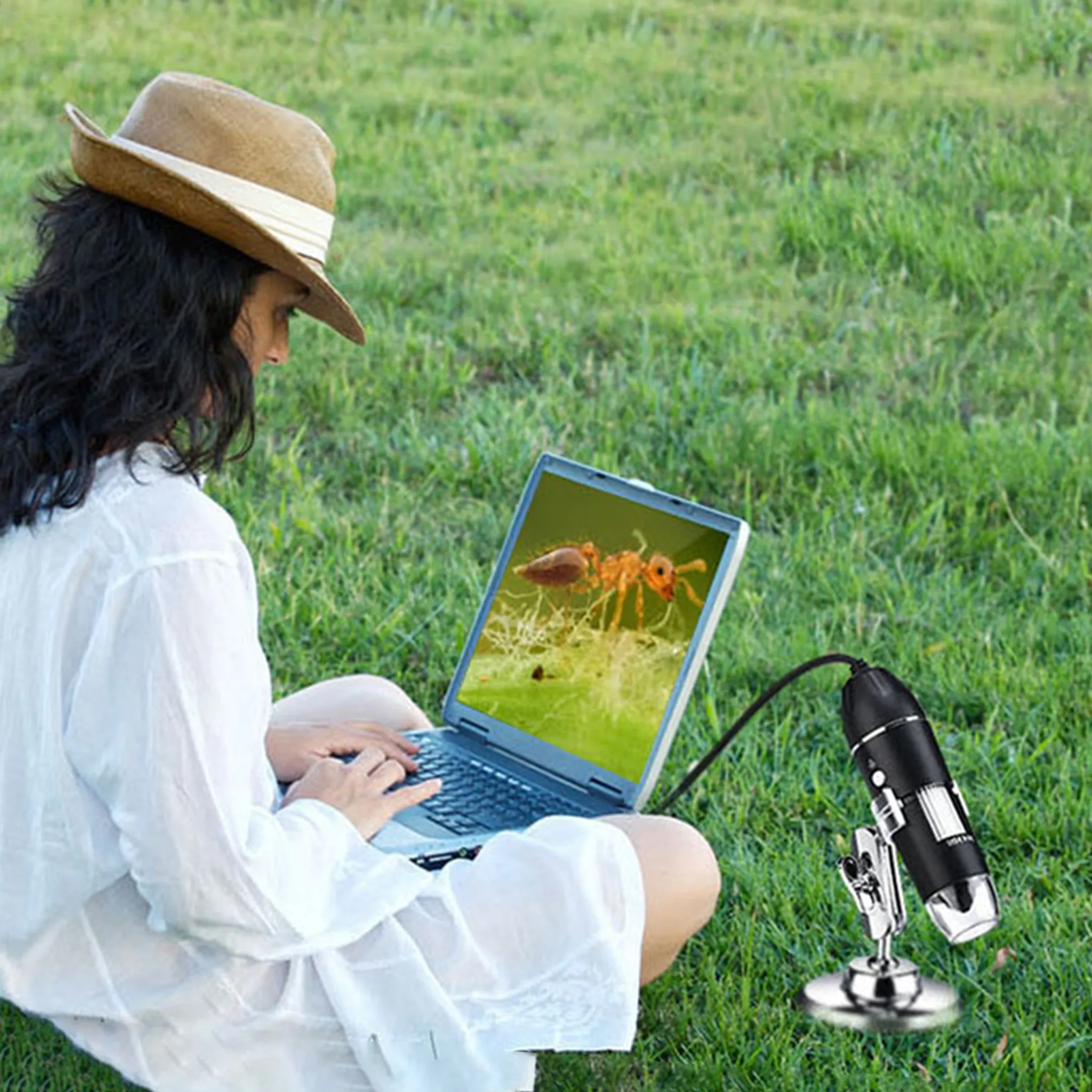 Eine Person mit Hut benutzt einen Laptop auf Gras. Der Bildschirm zeigt eine Ameise. Ein Mikroskop ist angeschlossen.