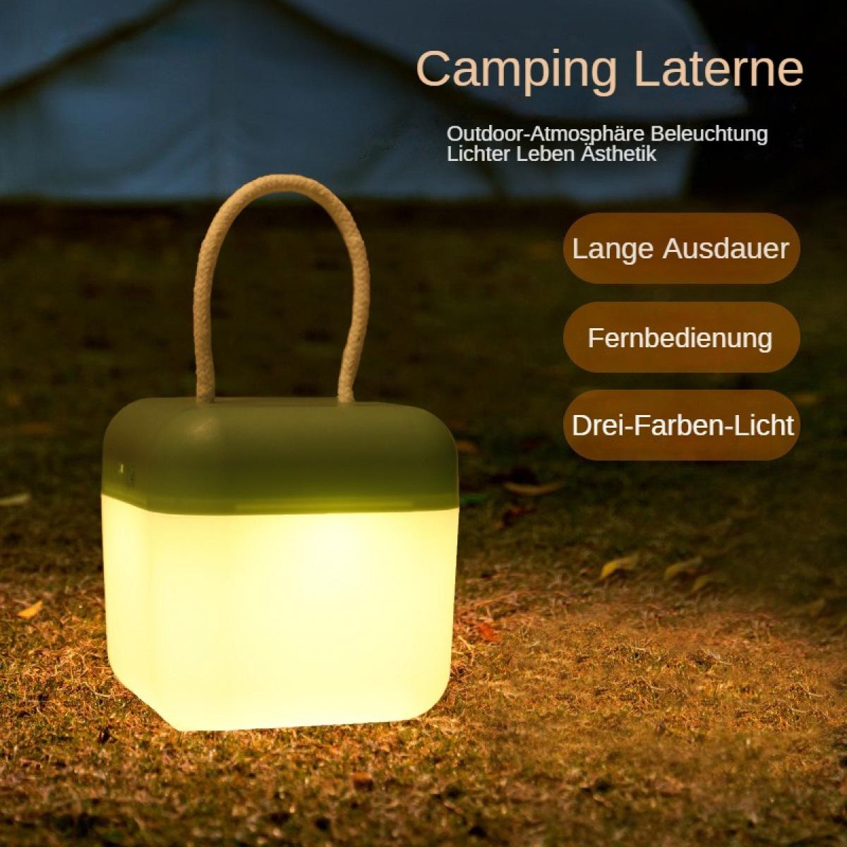 Eine leuchtende Campinglaterne mit Griff steht auf Gras. Die Oberseite ist grün, der Körper weiß, das Licht warm.