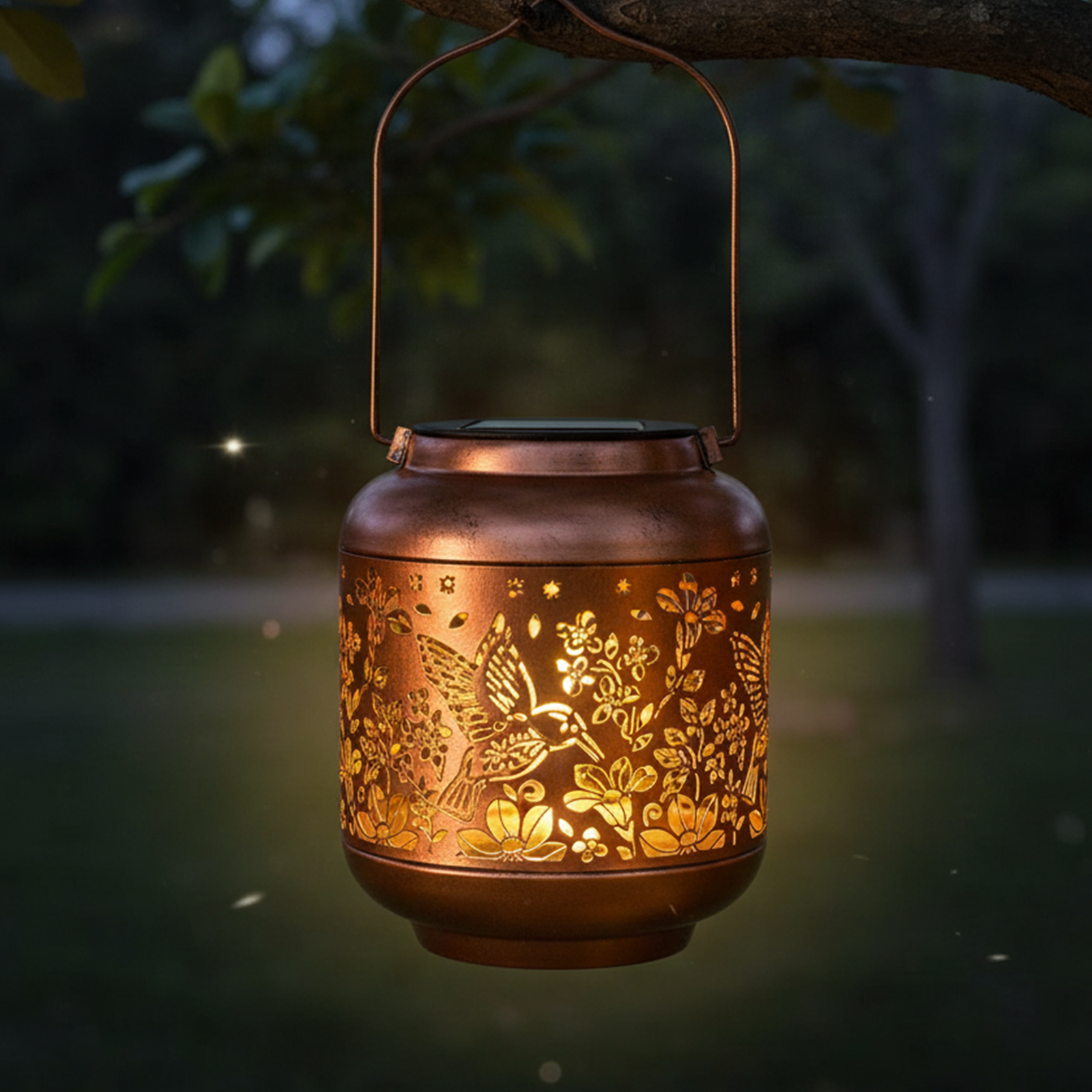 Eine bronzefarbene Laterne mit floralen und Kolibri-Ausschnitten hängt an einem Ast. Sie ist von innen beleuchtet, mit dunklem Hintergrund.