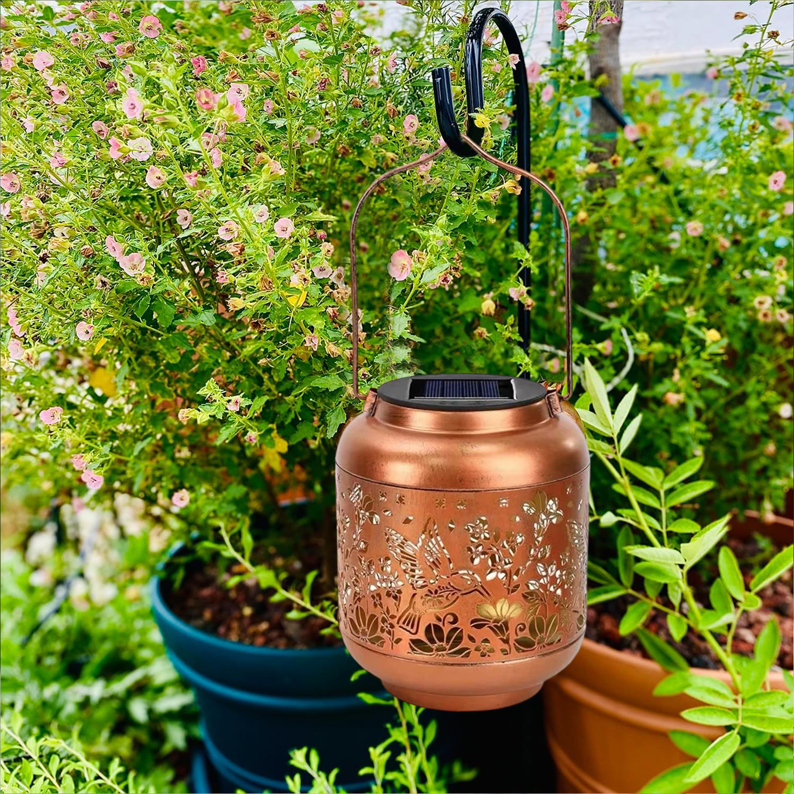 Eine Kupferlaterne mit Blumenausschnitten hängt in einem Garten, umgeben von Pflanzen und rosa Blüten.