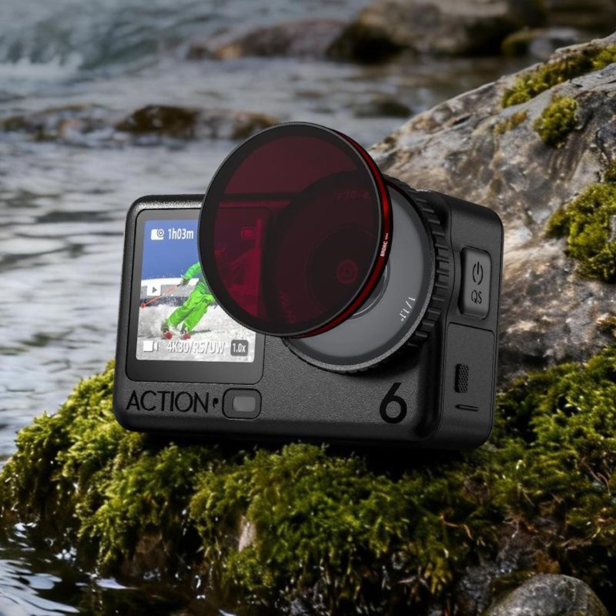 Schwarze Action-Kamera mit Rotfilter auf moosbewachsenem Felsen am Wasser. Bildschirm zeigt Skifahrer.
