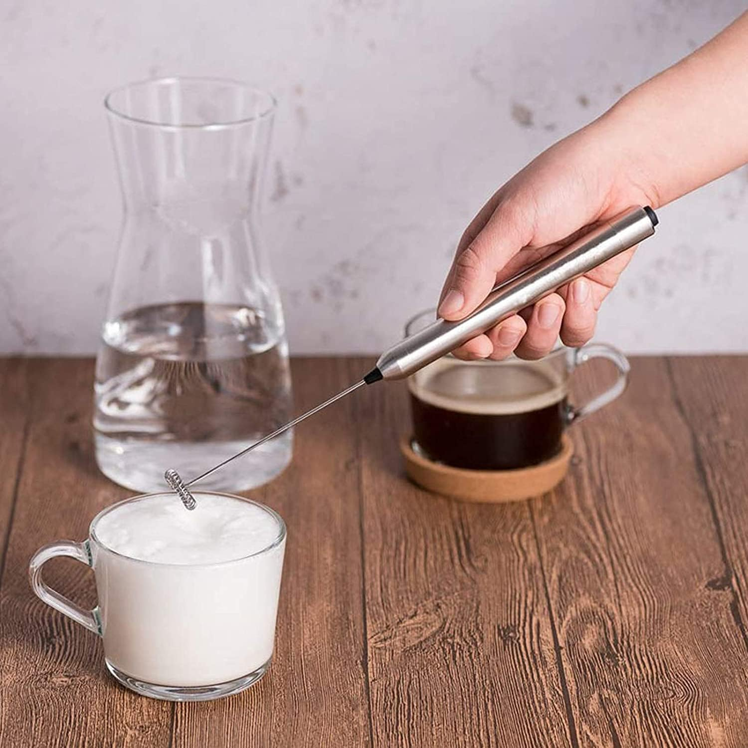 Eine Hand schäumt Milch in einer Glastasse mit einem Aufschäumer auf. Eine Kanne Wasser und eine Tasse Kaffee stehen daneben auf einem Holztisch.
