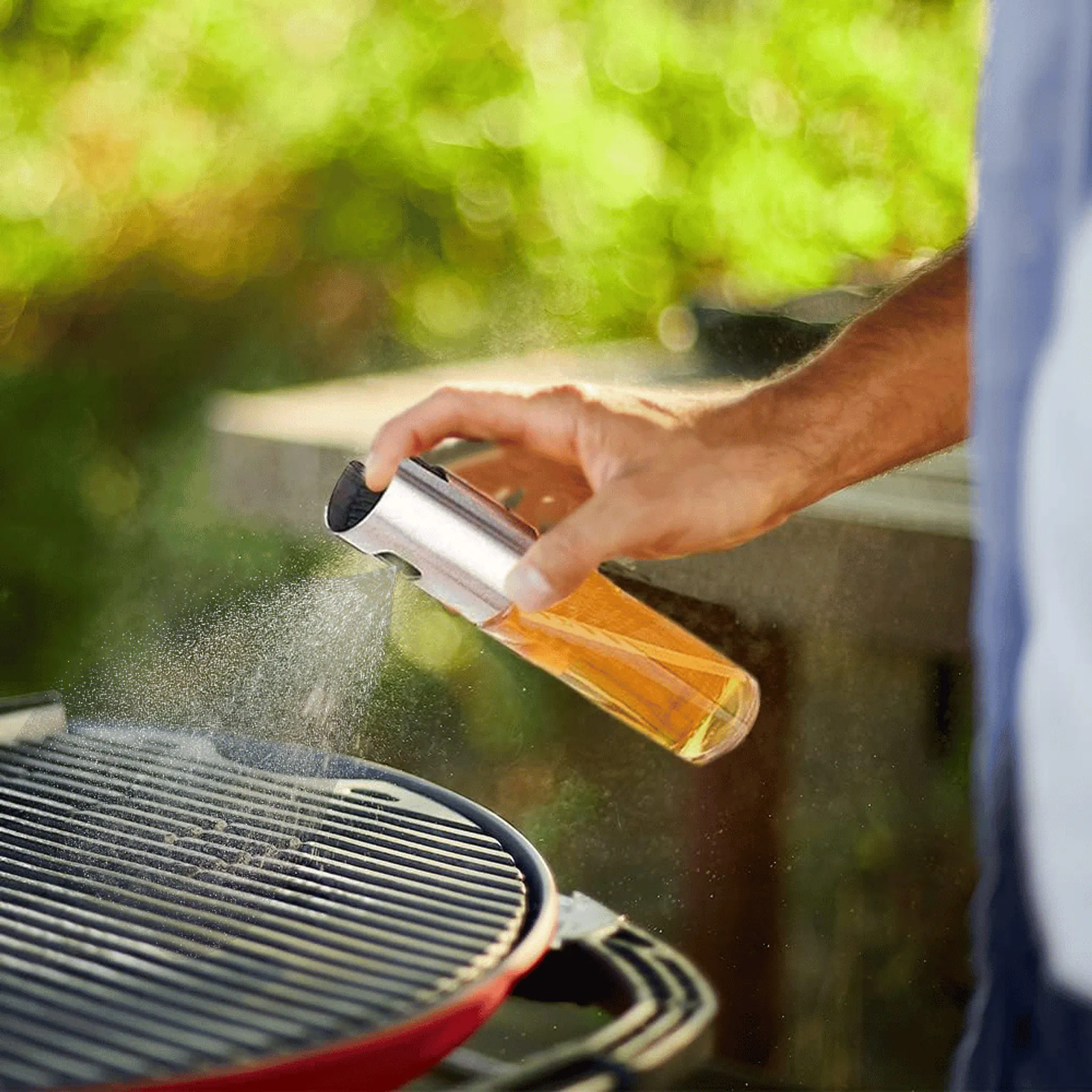 Eine Hand sprüht Öl auf einen Grill. Die Ölflasche ist silber und klar, der Grill ist schwarz und rot.